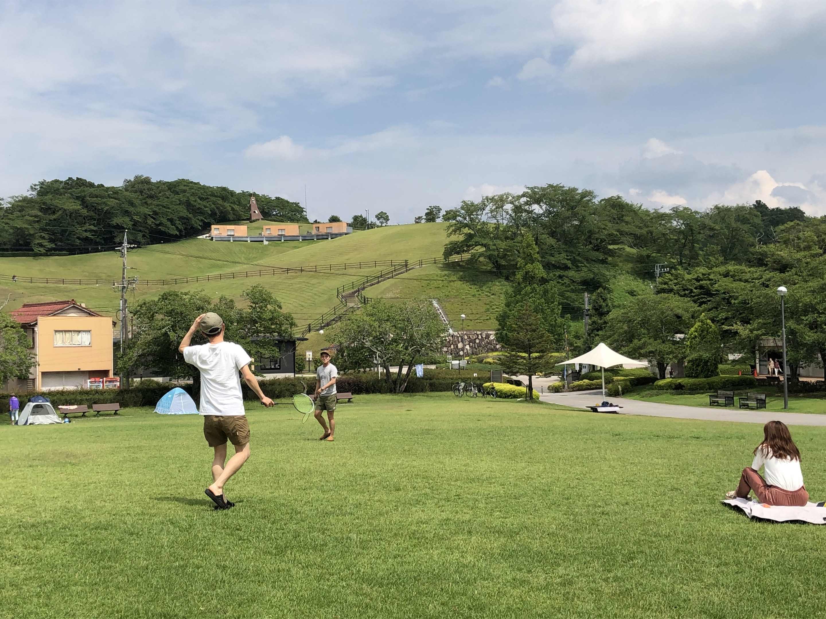 Eine Gruppe von Menschen, die Badminton in einem Park spielt, mit Zelten, Straßenlaternen und Gebäuden im Hintergrund.