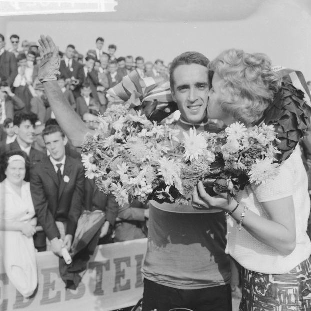 Ein Schwarz-Weiß-Foto von einem Mann und einer Frau, die sich in einer Menschenmenge umarmen und küssen, während der Mann einen Blumenstrauß hält und die Frau strahlt, während die Menge applaudiert.