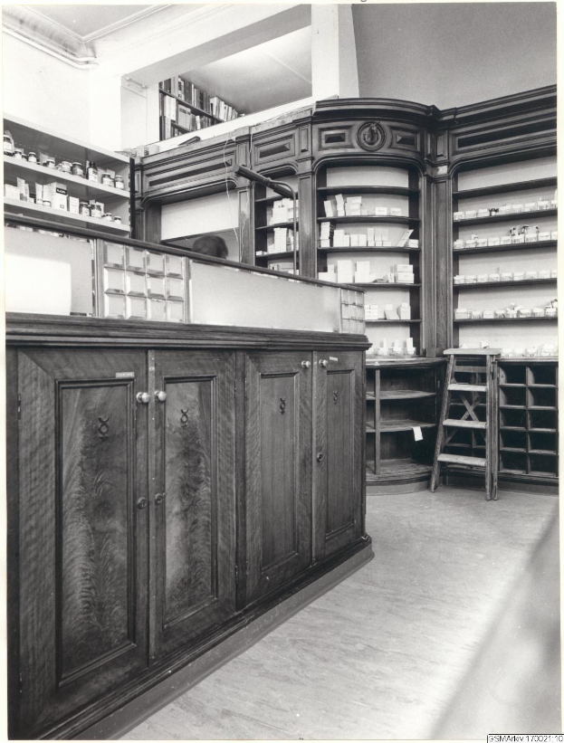 Schwarz-weiß-Foto eines vintage-Apothekeninneren mit einer Theke, Schüben, einer Leiter und Regalen, die mit verschiedenen Gegenständen bestückt sind, mit Text oben.