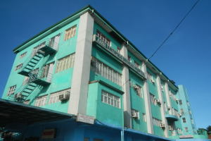 Ein große grünanlage mit zahlreichen Fenstern, Klimaanlagen, Treppen, Geländern, Namenstafeln, Strommasten, Stromkabeln, Kraftfahrzeugen auf der Straße, Bäumen und einem klaren blauen Himmel.
