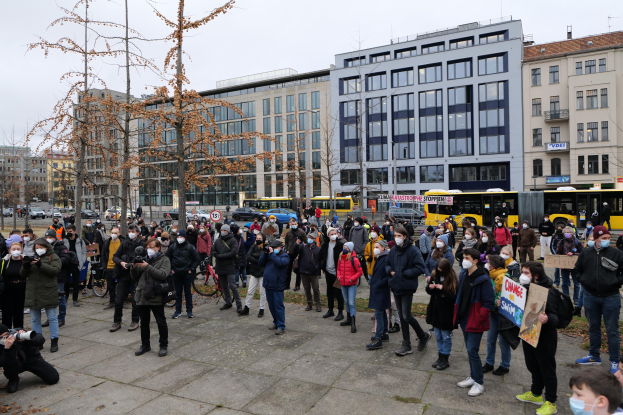 Eine große Gruppe von Menschen steht vor einem Gebäude während einer Demonstration in Berlin, einige tragen Masken und halten Schilder, mit Fahrzeugen, Bäumen und Gebäuden im Hintergrund.