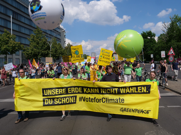 Eine Gruppe von Menschen geht eine Straße entlang und hält ein gelbes "Wähle für das Klima"-Schild hoch, mit Luftballons, Schildern, Fahrrädern, Bäumen, Laternenpfählen und Gebäuden im Hintergrund unter einem klaren blauen Himmel.