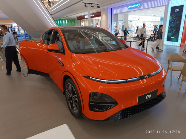 Orangefarbener Elektroauto in einer Ausstellungshalle umgeben von Menschen, Stühlen und einem Wagen rechts und Geschäften im Hintergrund.