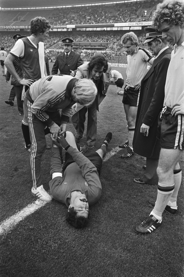 Schwarzes und weißes Bild eines Fußballspielers, der auf dem Boden liegt und von einer Gruppe von Männern umgeben ist, mit einer Stadionkulisse im Hintergrund; der Spieler scheint verletzt zu sein.