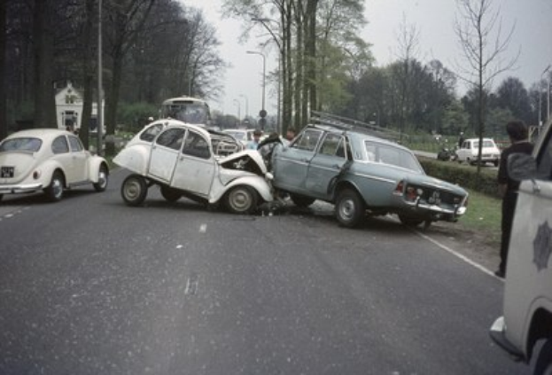 Zwei beschädigte Autos am Straßenrand mit Menschen in der Nähe, umgeben von Bäumen, Strommasten, Gebäuden und einem klaren blauen Himmel.