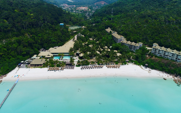 Luftaufnahme eines Strandresorts in Koh Samui, Thailand, mit einer Brücke links, Gebäuden, Bäumen, Hügeln und ein paar Menschen am Strand.