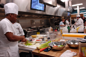 Eine Gruppe von Köchen in einer Küche, mit einem im Vordergrund in einer weißen Schürze und Mütze, der ein Messer hält und von Utensilien, Schüsseln, Gemüse, einem Handy, Flaschen und anderen Gegenständen umgeben ist, während im Hintergrund ein Fernseher und Deckenlampen zu sehen sind.