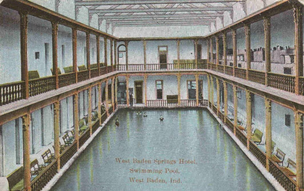 Schwarz-weiß-Postkarte eines Hallenbades im West Baden Springs Hotel mit schwimmenden Menschen, umgeben von Säulen, Geländern, Stühlen und anderen Gegenständen, mit Text unten.