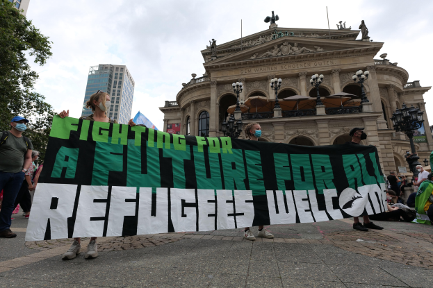 Eine Gruppe von Menschen mit Masken, die ein Transparent mit der Aufschrift "Kämpfen für eine Zukunft für alle Flühtlinge Willkommen" vor einem Gebäude mit Laternenmasten und Bäumen unter einem bewölktem Himmel halten.