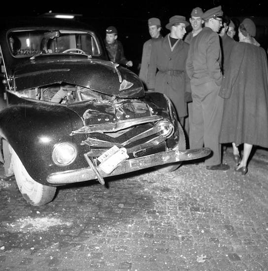 Schwarzes und weißes Bild eines schwer beschädigten Autos am Straßenrand mit eingedellter Motorhaube und zersplitterten Fenstern, umgeben von Menschen, die die Szene begutachten.