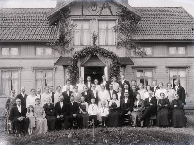 Schwarzes und weißes Foto einer großen Gruppe von Menschen, die vor einem Haus mit viel Grün und einem sichtbaren Himmel posieren.