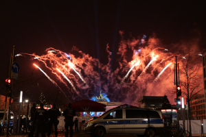 Ein Polizeiwagen steht vor einer Menschenmenge, mit beleuchteten Gebäuden, Bäumen, Pfählen, Verkehrszeichen, Schildern und Zelten im Hintergrund, unter einem Himmel voller bunter Feuerwerke.