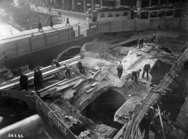 Schwarz-weiß-Foto von Bauarbeitern, die eine Brücke reparieren, mit Menschen auf der Brücke und Fahrzeugen im Hintergrund.