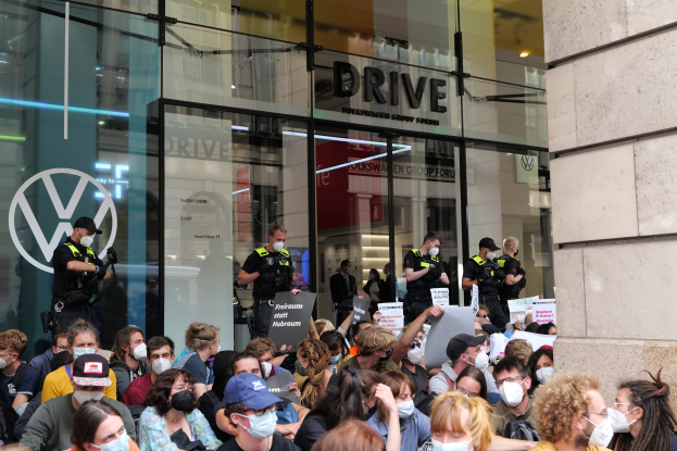 Eine große Gruppe von Menschen mit Masken und Schildern vor einem Gebäude mit Glaswänden und Text, einige tragen Mützen und halten Kameras, was auf eine Demonstration in Melbourne, Australien, hinweist.