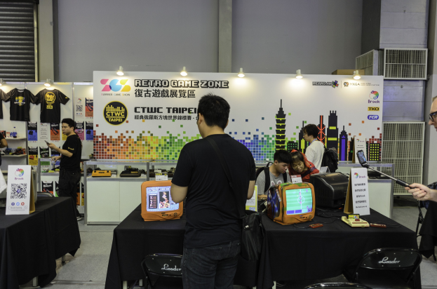 Leute versammeln sich um einen Tisch mit einem Videospiel, mit Stühlen, einer Wand, aufgehängten T-Shirts und Deckenbeleuchtung im Hintergrund.