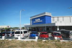 Ein Autohaus mit vorne geparkten Fahrzeugen, umgeben von einem Metallzaun, Gras, Pflanzen, Bäumen, Straßenlaternen und einem bewölkten Himmel im Hintergrund.