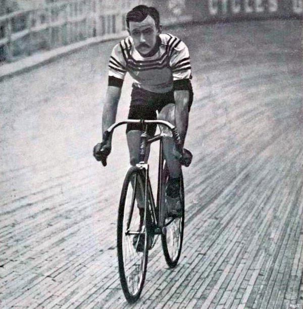 Schwarz-weiß-Foto eines Mannes, der auf einer Bahn einen Fahrrad fährt, mit einem Zaun und einer Tafel im Hintergrund.