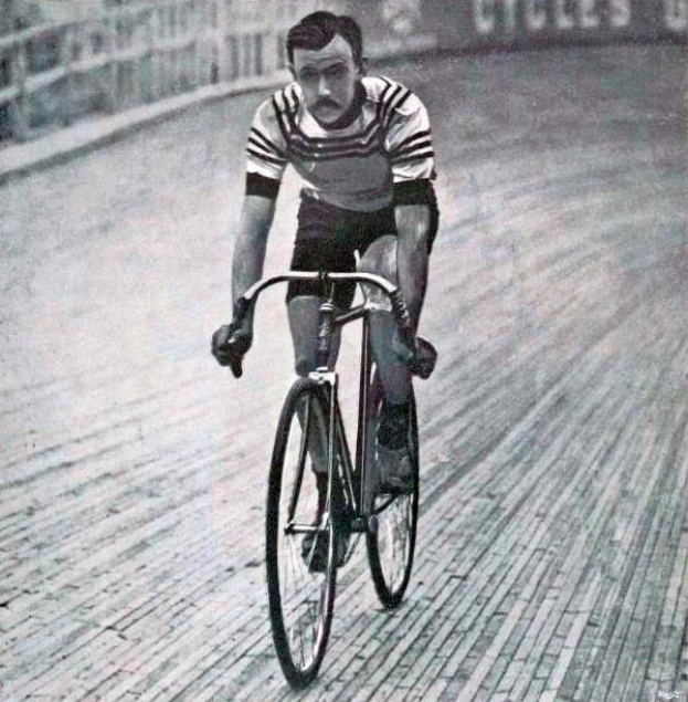 Schwarz-weiß-Foto eines Mannes, der auf einer Bahn einen Fahrrad fährt, mit einem Zaun und einer Tafel im Hintergrund.