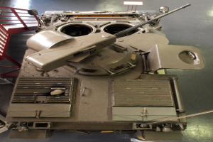 Ein großer Militärpanzer mit sichtbaren Ketten und Turm steht auf einem Boden, mit einem Metallgeländer auf der linken Seite.