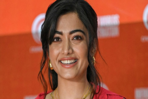 Eine Frau in einem roten Kleid mit einem eleganten Dutt und dezentem Make-up lächelt in die Kamera, trägt schwarze High Heels und hat einen hellgelben Banner mit der Aufschrift "Sonam Kapoors beste Beauty-Looks vom Cannes Film Festival" im Hintergrund.