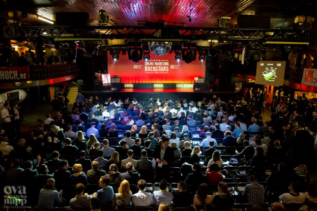 Eine große Menschenmenge sitzt auf Stühlen vor einer Bühne mit Bildschirmen und Bannern, beleuchtet von Scheinwerfern und umgeben von Geländern, voller Erwartung auf den Beginn eines Online-Marketing-Rockstars-Events, mit einem Wasserzeichen in der linken unteren Ecke.