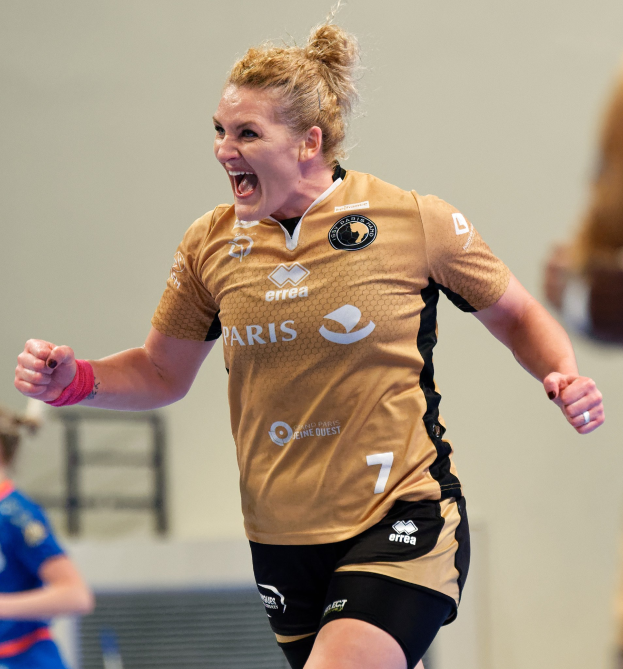 Eine Frau in brauner Bluse und schwarzen Shorts rennt auf einem Futsalplatz, feiert mit triumphierendem Gesichtsausdruck, während zwei andere Frauen im Hintergrund sichtbar sind.