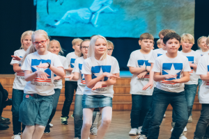 Eine Gruppe von Kindern in weißen Hemden und blauen Shorts, einige mit Brille, tanzt auf einer Bühne mit Zuschauern links und einem Bildschirm im Hintergrund und nimmt an einem Tanzwettbewerb teil.