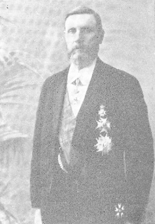 Ein Mann in einem schwarzen und weißen Anzug mit einem weißen Hemd und schwarzer Krawatte steht vor einer üppigen grünen Pflanze, Gesichtsausdruck ernst.