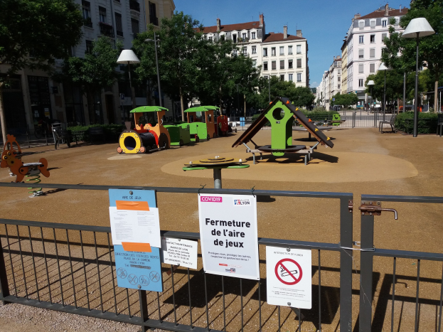 Ein umzäunter Spielplatz in einem urbanen Platz mit Schildern am Zaun, umgeben von Bäumen, Laternenmasten, Gebäuden und einem sichtbaren Himmel.