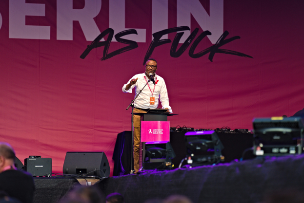 Ein Mann in einem weißen Hemd und braunen Hosen steht auf einer Bühne und hält ein Mikrofon in der Hand, hinter ihm eine pinkfarbene Fahne mit der Aufschrift 'Berlin as Fuck'.