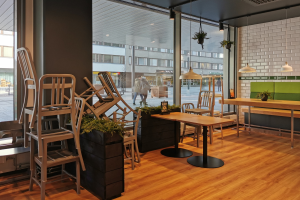 Ein Restaurant-Innenraum mit Holztischen und -stühlen, großen Fenstern mit Blick auf eine Stadtstraße mit Gebäuden, Pfählen und Menschen, umgeben von Pflanzen und Lichtern.