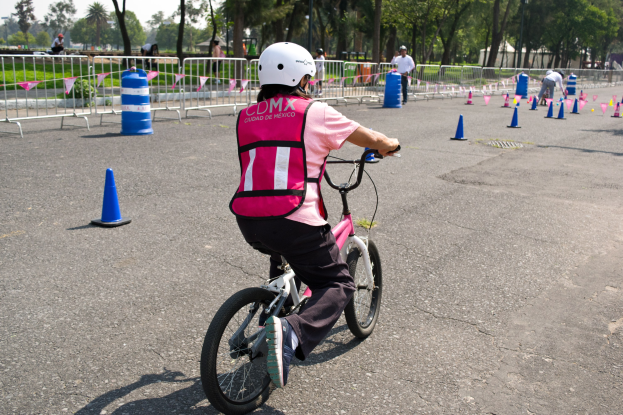 Eine Person in pinker und schwarzer Kleidung mit einem weißen Helm fährt auf einem Fahrrad eine von Verkehrskegeln gesäumte Straße entlang, mit einigen stehenden Menschen, einem Geländer, Bäumen und einem blauen und weißen Himmel im Hintergrund.