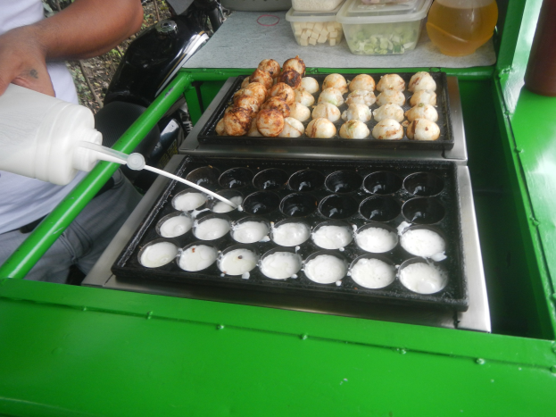 Ein Mann gießt Milch aus einer Flasche in eine Schale mit Essen auf einem grünen Tisch, umgeben von anderen Schalen, Flaschen und Gegenständen.