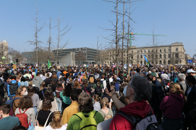 Große Menschenmenge mit Schildern und Taschen vor einem Gebäude mit Bäumen und klarem Himmel, was auf eine Klimaprotestaktion hindeutet.