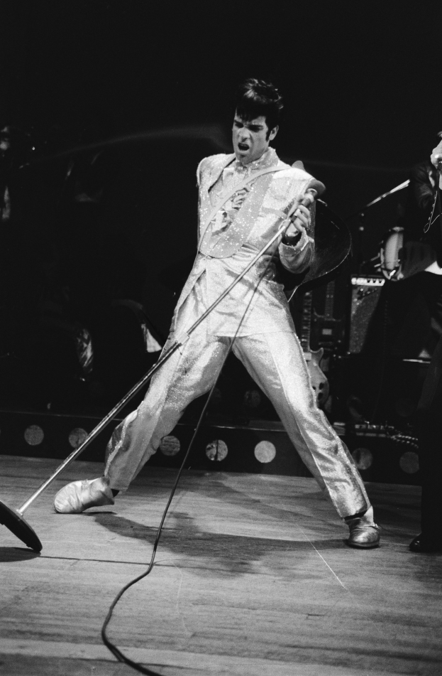 Elvis Presley in weißer Jumpsuit performs auf der Bühne im Apollo Theater in Las Vegas, hält ein Mikrofon in der Hand und hat rechts neben sich einen Musiker mit einem Instrument; Bühnenlichter und Musikinstrumente sind im Hintergrund zu sehen.