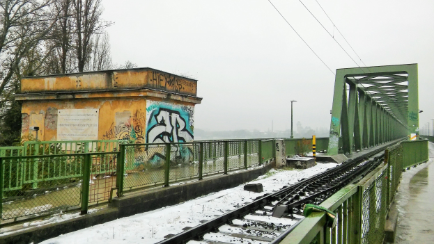 Eisenbahnschiene mit Graffiti an der Seite, umgeben von Geländern, Pfählen, Lampen, Drähten und Bäumen, mit schneebedeckter Erde und sichtbarem Himmel im Hintergrund.