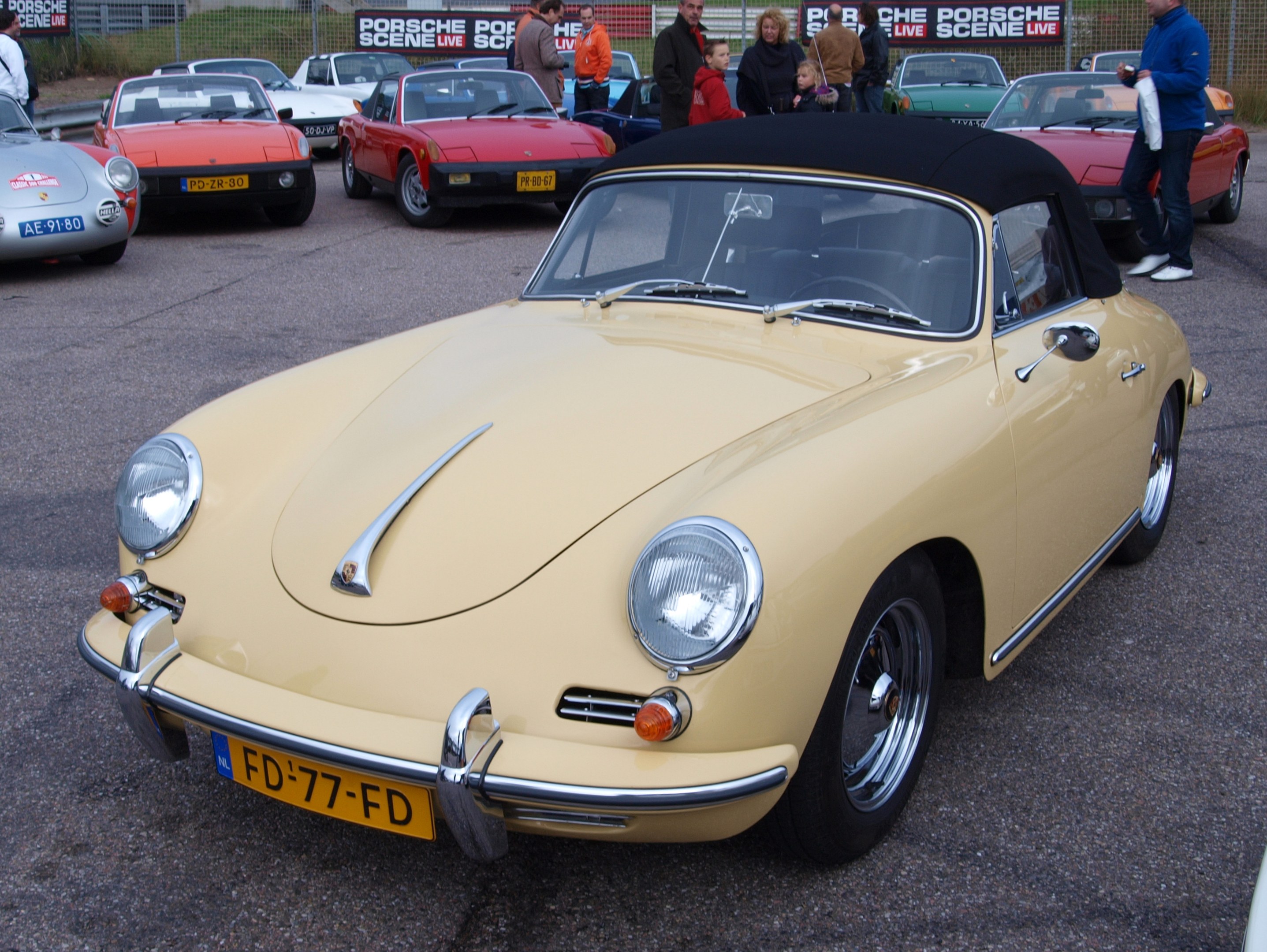 Ein gelber Porsche 356 Speedster auf einem Parkplatz mit Menschen in der Nähe, umgeben von einem Zaun, Bannern, Bäumen, Gebäuden und einem klaren Himmel.