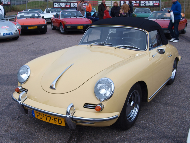 Ein gelber Porsche 356 Speedster auf einem Parkplatz mit Menschen in der Nähe, umgeben von einem Zaun, Bannern, Bäumen, Gebäuden und einem klaren Himmel.