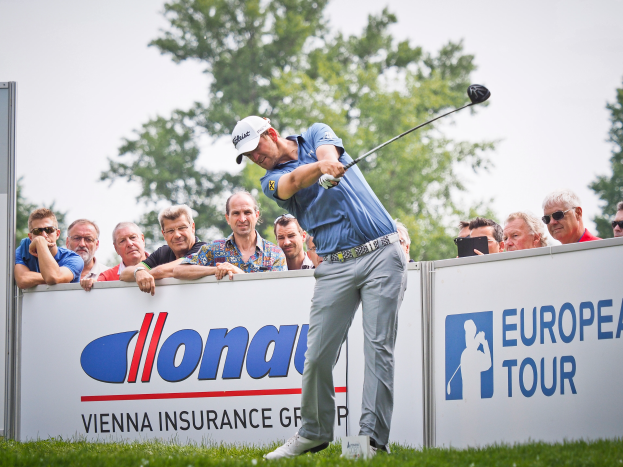 Ein Mann mit Mütze schwingt einen Golfschläger auf einem grünen Feld, mit Text- und Logo-Tafeln, Bäumen und einem klaren blauen Himmel im Hintergrund.