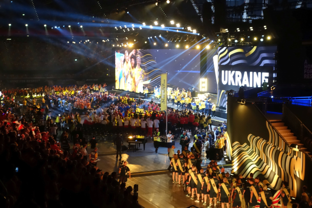 Eine große Menschenmenge auf einer hell erleuchteten Bühne mit Musikern, die Instrumente spielen, und einem Bildschirm im Hintergrund, der auf ein bevorstehendes Ereignis hindeutet.