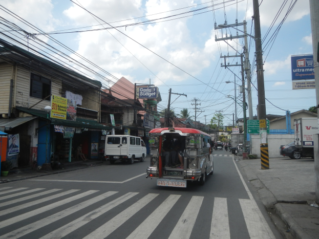 Eine Stadtstraße mit Fahrzeugen, Strommasten, Gebäuden, Schildern, Bäumen und einem bewölkten Himmel, mit einem Bus, der an hohen Gebäuden vorbeifährt.