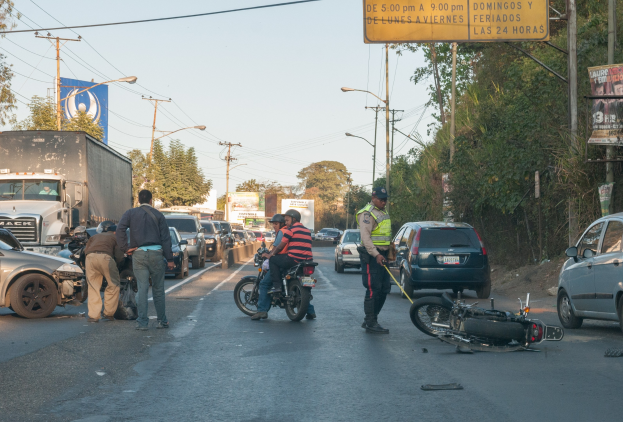 Gruppe von Menschen um ein verunglütztes Motorrad auf der Straße mit mehreren Fahrzeugen, darunter ein Lastwagen, und Hintergrundelementen wie Bäume, Masten, Lichter, Schilder und Himmel.