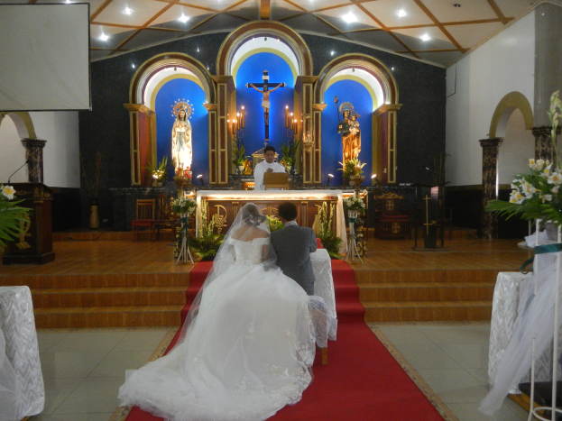 Ein Brautpaar sitzt auf einem roten Teppich vor einem Kirchenaltar, flankiert von Blumenbouquets, mit Statuen, Kerzen, Stühlen, einem Podium, einem Bildschirm und Deckenlampen im Hintergrund.