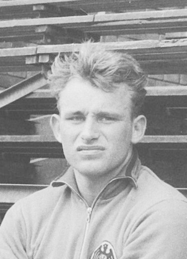 Schwarzes und weißes Foto eines älteren Mannes in einer Jacke, der vor gestapelten Holzplanken steht, der als legendärer deutscher Fußballspieler gilt.