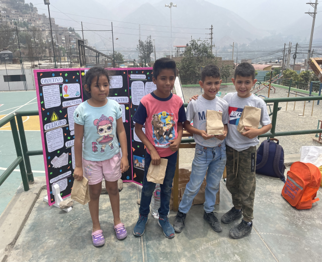 Gruppe von Kindern auf einem Basketballplatz mit Papiertüten in den Händen, mit Gegenständen auf der rechten Seite und einer Tafel mit Text auf der linken Seite, vor einem Hintergrund aus Zaun, Pfosten, Straßenlaternen, Bäumen, Gebäuden, Hügeln und Himmel.