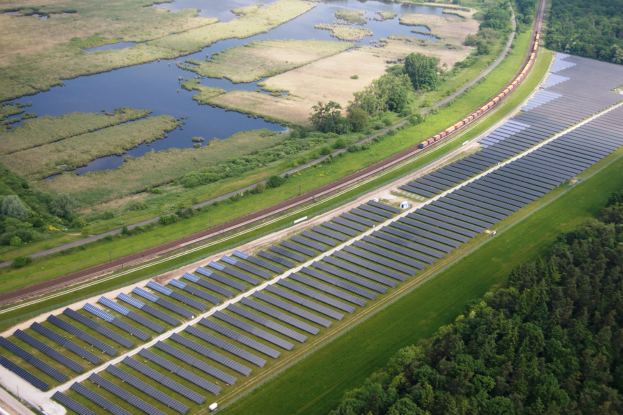 Luftbild einer Solarpark mit Solarpanelen in einem Feld, umgeben von Bäumen, Gras, Pflanzen und Wasser, mit einem Zug auf einer nahen Bahnstrecke.