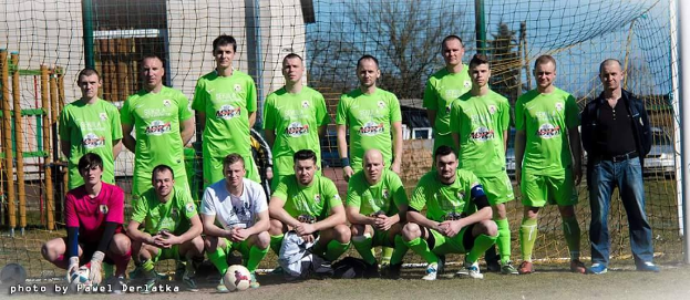 Eine Gruppe von Männern in grünen T-Shirts steht und hockt auf einem Fußballfeld mit einem Ball vor ihnen, ein Torpfosten rechts und ein Hintergrund aus Bäumen, Gebäuden, Pfählen, Fahrzeugen und Himmel, mit einer Reihe von Zahlen links unten.
