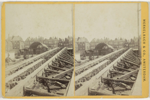 Schwarz-weiß-Foto einer Zugschiene mit Gebäuden im Hintergrund, Text 'Merkelbach & Co, Co. Amsterdam' auf der rechten Seite und verstreute hölzerne Gegenstände im Vordergrund.