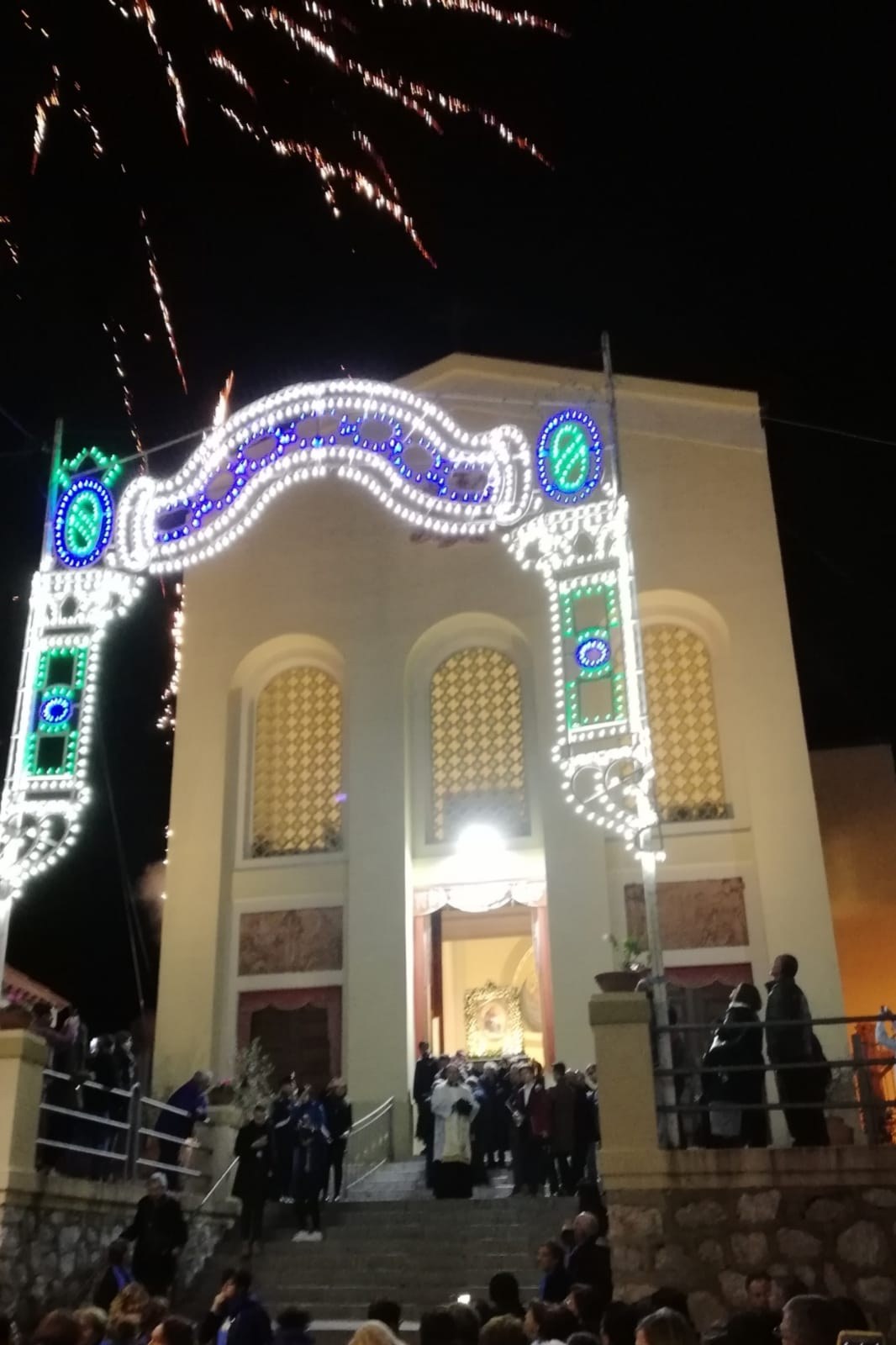 Eine Gruppe von Menschen steht vor einem Gebäude mit einem bogenförmigen, mit Lichtern geschmückten Eingang, Treppen mit Geländern und Topfpflanzen, während Feuerwerke den Nachthimmel erhellen, während sie das neue Jahr feiern.