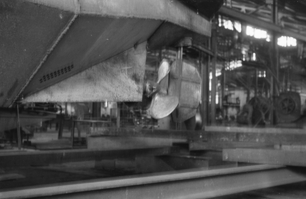 Ein Schwarz-Weiß-Foto eines Boots in der Fertigung in einer Fabrik, umgeben von metallenen Objekten, Pfählen und Lichtern.